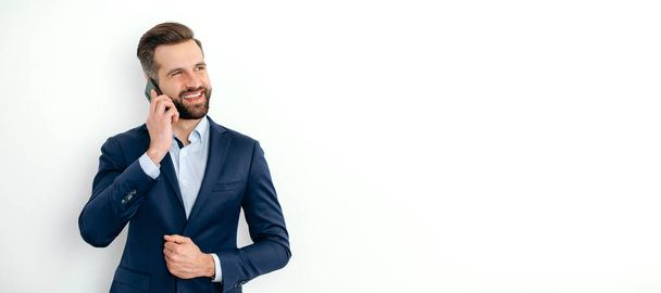 Handygespräche. Panoramafoto eines kaukasischen Geschäftsmannes im eleganten Anzug, steht auf einem weißen, isolierten Hintergrund, spricht auf dem Smartphone mit einem Kollegen, schaut weg, lächelt. Kopierraum - Foto, Bild