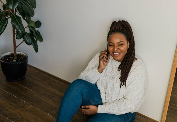 Positive young african american woman chatting on cellphone and smiling on floor at home. Cheerful young woman communicating with someone on smartphone - Photo, Image