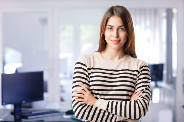 Confident young businesswoman wearing casual clothes and standing with arms crossed at the office. - Foto, imagen