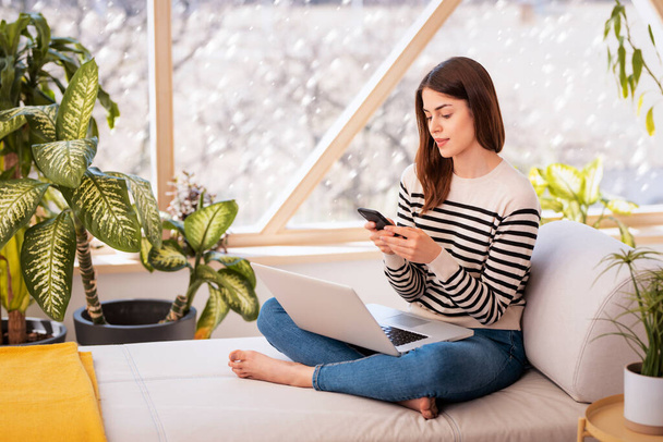Volledige lengte van jonge vrouw sms-en en met behulp van laptop terwijl zitten op de bank thuis in de winter tijd. - Foto, afbeelding