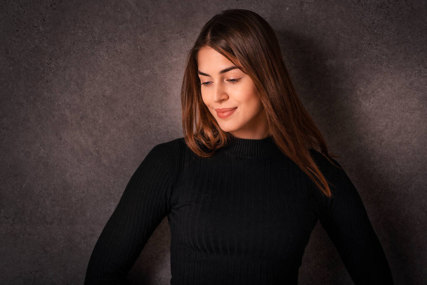 Portrait of smiling young woman standing at isolated background. Confident young female is having brunette hair. She is in black sweater against dark background. Copy space.  - Valokuva, kuva