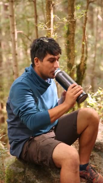 A young latino man drinking water from a bottle in the forest. Small break during hiking in the mountains. Healthy lifestyle and activity to enjoy adventures in nature concept in vertical 4K - Footage, Video