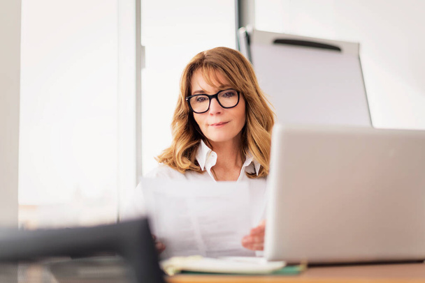 Mid aged professional woman wearing shirt and using laptop while working in a modern office. Attractive businesswoman sitting at office desk. - Foto, imagen