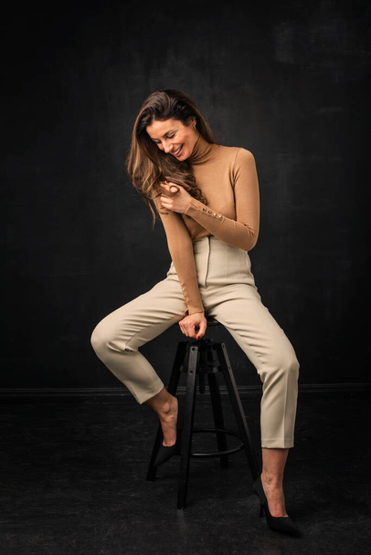 Full length of an attractive woman with toothy smile sitting at isolated black background. Brunette haired female wearing turtleneck sweater and trousers while sitting against isolated dark background. Copy space.  - Photo, image