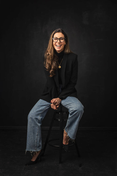 Full length of an attractive woman toothy smiling and looking at camera. Brunette haired female wearing blazer and eyewear while sitting against isolated black background. Copy space.  - Fotoğraf, Görsel