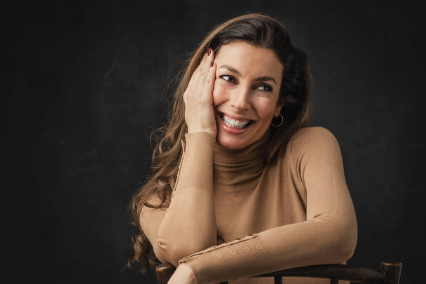 Studio portrait of an attractive middle aged woman with toothy smile wearing turtleneck sweater while sitting at isolated dark background. Copy space. - Foto, afbeelding