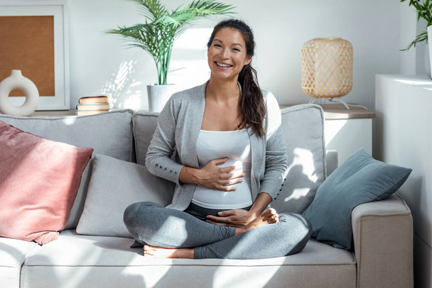 Shot of beautiful pregnant woman touching her belly sitting in lotus position on couch in living room at home. - 写真・画像