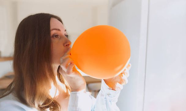 Portrait of young woman blowing orange balloon, getting ready to celebrate birthday. Home decorations for party. Woman in living room on daytime. Concept of emotions, lifestyle - Fotoğraf, Görsel