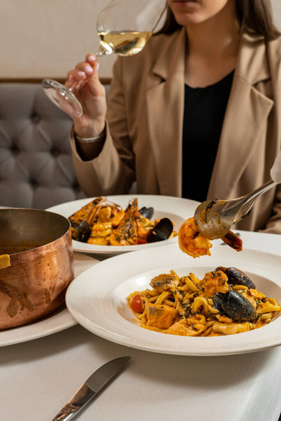 Homemade pasta with shrimp, salmon, mussels in a wine-cream sauce in a copper dish with a handle and baked in the oven under a dough lid. Place the dishes on a table with a light tablecloth. The waiter uses a fork and a spoon to place pasta and white - Φωτογραφία, εικόνα