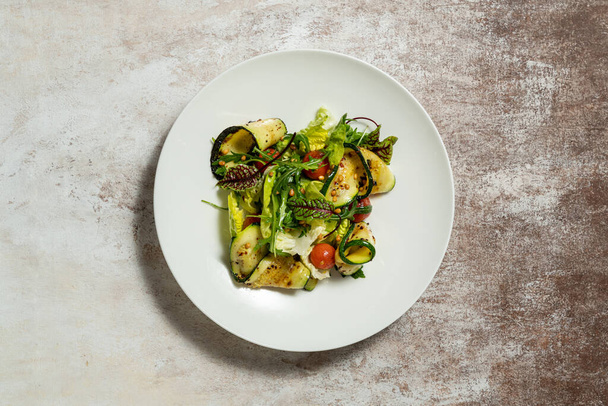 Gemüsemischsalat mit Zucchini, Kirschtomaten, Pinienkernen, Olivenöl und Balsamico-Soße. Essen liegt in einem hellen Keramikteller auf farbigem Hintergrund. - Foto, Bild