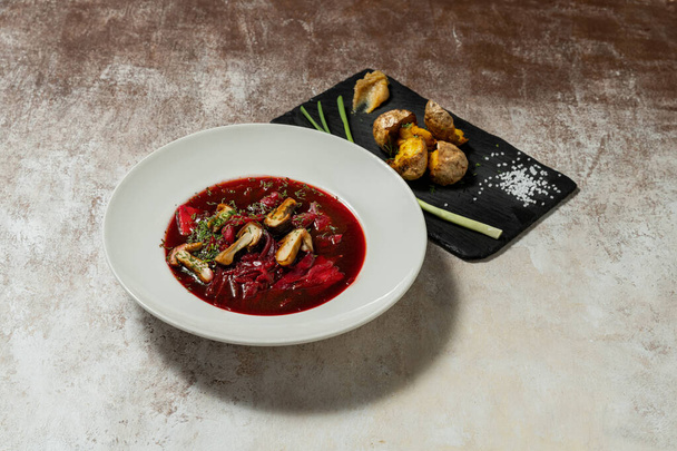 Ukrán böjt leves borscht, gombával és zöldséggel egy könnyű kerámia tányéron. A közelben van egy palatábla sült krumplival és zöldhagymával. Ételek állni egy könnyű szövet háttér. - Fotó, kép