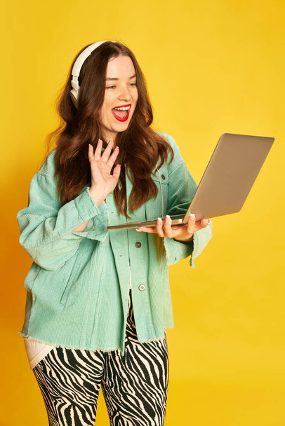 Lección en línea, estudio. Una joven estudiante, una chica bonita con auriculares sosteniendo la computadora portátil y saludando a la pantalla sobre el fondo amarillo del estudio. Concepto de educación, moda, estilo de vida, juventud - Foto, Imagen