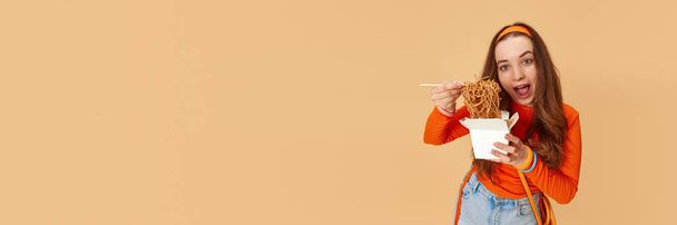 Nourriture pourrie et malsaine. Bannière avec jeune fille adorable tenant boîte avec de la nourriture et de manger des nouilles avec visage affamé sur fond de studio de gingembre. Concept de suralimentation, alimentation, restrictions, sport, publicité - Photo, image