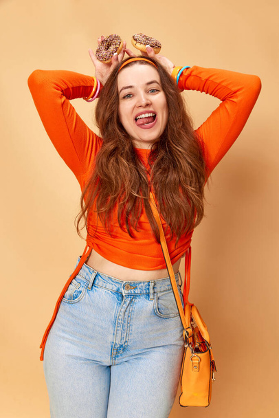 Funny bunny. Portrait of happy, beautiful girl wearing bright clothes, jeans posing with donuts in hands and smiling on ginger background. Concept of fashion, beauty, lifestyle, human emotions, ad - Photo, Image