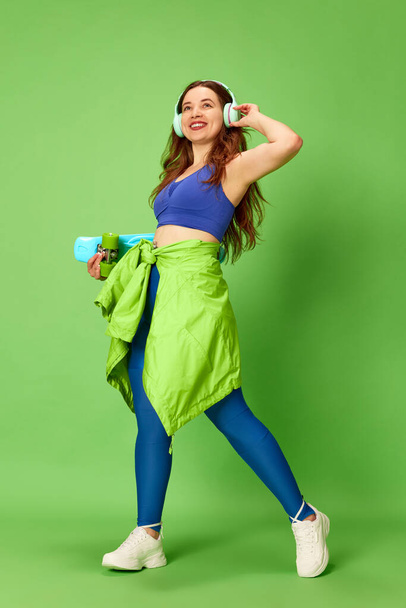 Jonge skateboarder. Portret van een mooi meisje met sportkleding en een koptelefoon met een skate en een glimlach op een groene studio achtergrond. Begrip hobby, sport, activiteit, jeugd, menselijke emoties, reclame - Foto, afbeelding