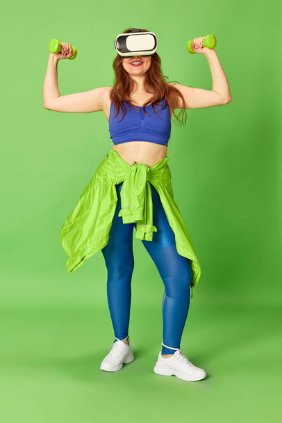 Girls power. One young charming girl, woman wearing vr glasses doing workout with dumbbells and smiling at camera over green background. Sport, fitness, healthy, fashion, activity concept - Фото, зображення
