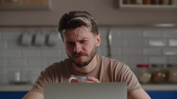 Een man zit aan een tafel met een kop koffie in zijn handen en kijkt zorgvuldig naar iets op een laptopscherm. Een fronsende man luistert naar een online lezing en drinkt koffie uit een kopje. Hoge kwaliteit 4k - Video