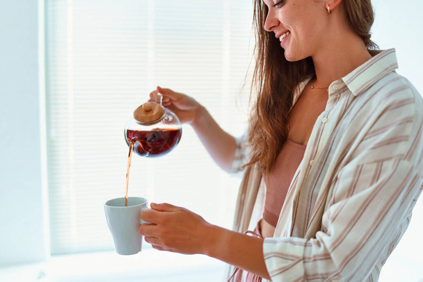 Happy young cute joyful smiling satisfied woman pouring hot tea into cup from glass teapot at home - Фото, зображення