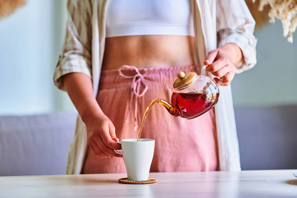 Woman pouring hot aromatic black tea into cup from glass teapot at home - Foto, Bild