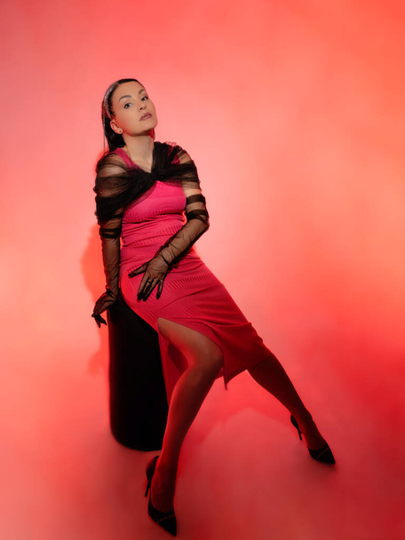 Portrait of a beautiful brunette in a red dress on a pink background. She is wearing elegant black gloves. - Photo, Image