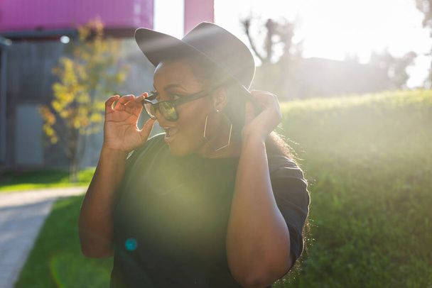Heureuse jeune femme afro-américaine portant un t-shirt riant en regardant la caméra debout dans la rue. génération z hipster fille posant extérieur rétroéclairé avec portrait de la lumière du soleil - Photo, image