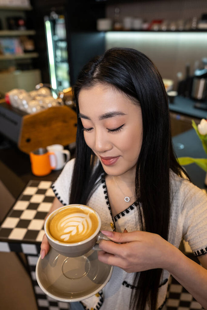 Smiling contented attractive long-haired Asian female looking at foam on the surface of her cappuccino - Fotó, kép
