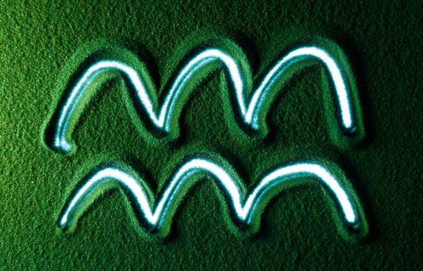 Yeşil Kum 'da Kova burcunun sembolünü elle çizmek. Erkek el, Kova burcunun sembolünü yeşil kumun üzerine beyaz arka ışıklandırmalı olarak yazar. Üst görünüm 4k çözünürlüğü - Fotoğraf, Görsel