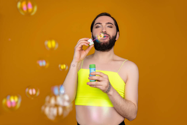 Playful bearded transsexual person wearing yellow top blowing soap bubbles having fun posing isolated over orange background - Fotó, kép