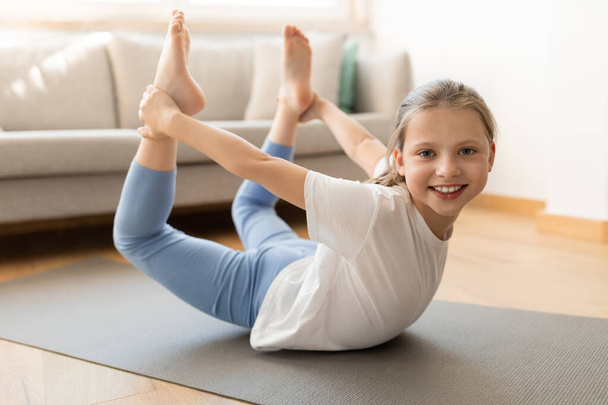 Veselý pěkný evropský dospívající dívka ve sportovním oblečení dělá pilates cvičení pro nohy v obývacím pokoji interiéru, zblízka, plné délky. Sport, léčba, péče o tělo a fitness doma - Fotografie, Obrázek