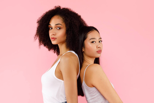 Portré fiatal, csinos ázsiai és afro-amerikai nőkről, akik háttal állnak egymásnak, együtt pózolnak és rózsaszín háttérrel néznek a kamerába. Életmód sokszínű nemzetiség emberek koncepció - Fotó, kép