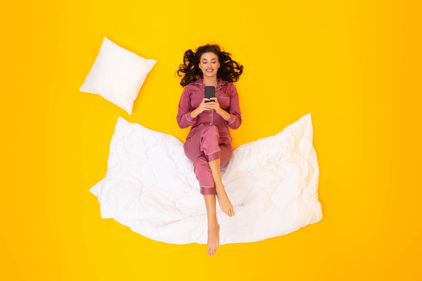 Ευτυχισμένη κυρία χρησιμοποιώντας το κινητό εφαρμογή στο τηλέφωνο που βρίσκεται στο Κουβέρτα φορώντας πιτζάμες πάνω από κίτρινο φόντο στούντιο. Γυναίκα γραπτών μηνυμάτων και επικοινωνίας στο διαδίκτυο πριν από τον ύπνο. Άνω όψη - Φωτογραφία, εικόνα