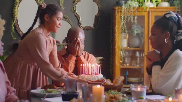 Afrikkalainen amerikkalainen mies sulkee silmänsä käsillään, kun tytär tuo syntymäpäiväkakkua, puhaltaa kynttilöitä vieraina hymyillen ja taputtaen käsiään kotiin perheillallinen. - Materiaali, video