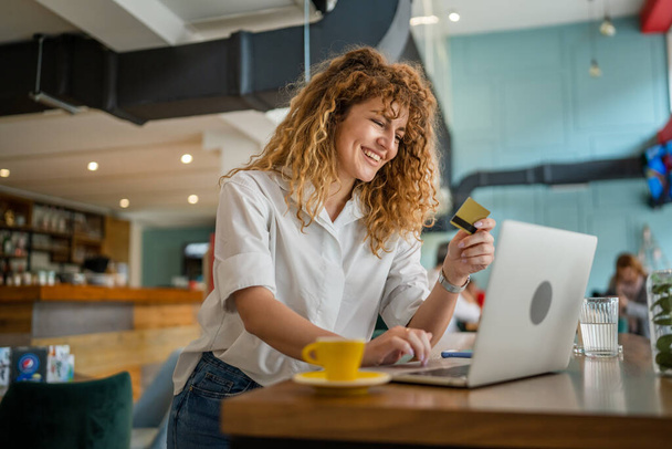 Jedna kobieta dorosła kaukaska kobieta z kręconymi włosami szczęśliwy uśmiech użyć karty kredytowej zakupy online na laptopie komputera w kawiarni prawdziwi ludzie e-bankowość sprzedaż internetowa zakup przestrzeni pozytywnych emocji - Zdjęcie, obraz