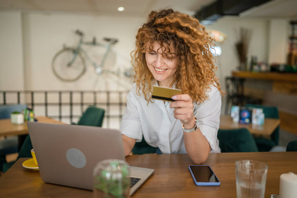 Одна женщина взрослая белая женщина с вьющимися волосами счастливая улыбка использовать кредитные карты покупки онлайн на ноутбуке компьютер в кафе реальные люди электронного банкинга интернет-продажи купить копия пространства положительные эмоции - Фото, изображение