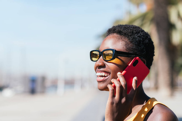 Vrolijke Afrikaanse vrouw die buiten telefoneert op een zonnige dag. Ze draagt casual zomerkleding en glimlacht gelukkig - Foto, afbeelding
