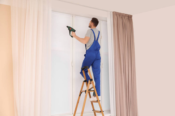 Worker in uniform installing roller window blind on stepladder indoors - Foto, afbeelding