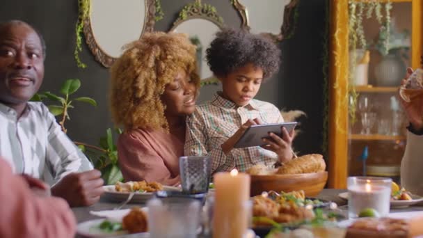 Pequeño niño afroamericano sentado en las rodillas de la madre cariñosa, sonriendo y jugando en la tableta digital durante la cena familiar de vacaciones en casa - Imágenes, Vídeo