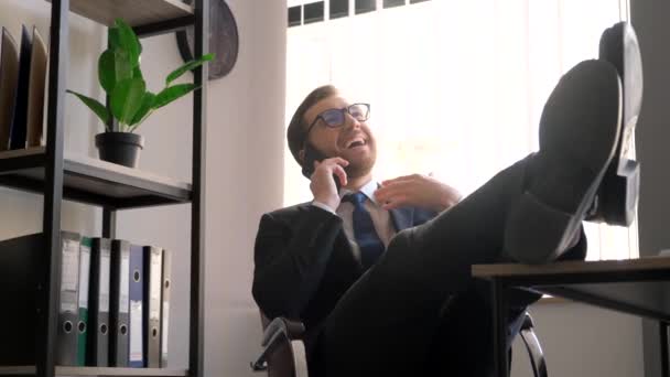 Ein zufriedener Geschäftsmann am Arbeitsplatz hat die Füße auf den Tisch gelegt und spricht mit dem Smartphone. Selbstbewusster und trotziger Büroangestellter hinter einem Laptop legte seine Füße auf den Tisch. - Filmmaterial, Video