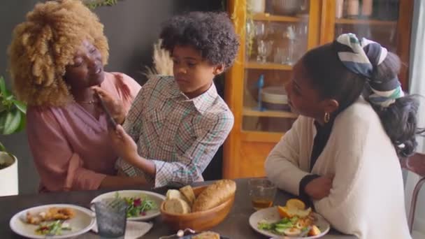 Mały Afroamerykanin siedzi na kolanach wesołej matki i pokazuje coś na tablecie cyfrowym rodzinie podczas domowego obiadu - Materiał filmowy, wideo