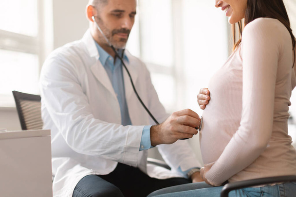 Male middle aged gynecologist with stethoscope examining belly of pregnant female patient, listening babys heartbeat while making check up to pregnant woman in clinic - Foto, Imagem
