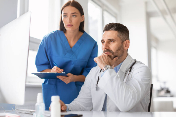 Összpontosított két orvos nézi a számítógép képernyőjén, megvitatják a betegek betegségek vagy diagnózis, együtt dolgoznak a klinikán. Középkorú férfi háziorvost segítő fiatal nővér - Fotó, kép