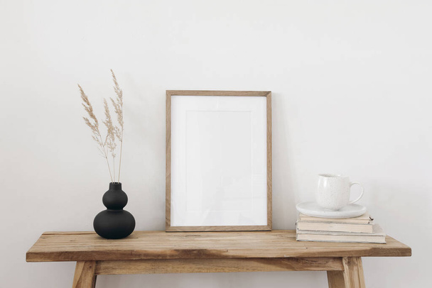 Blank vertical wooden picture frame mockup. Organic shaped black vase with dry grass on table, bench. Cup of coffee on old books. Working space, home office., art, poster display. Boho beige interior. - 写真・画像