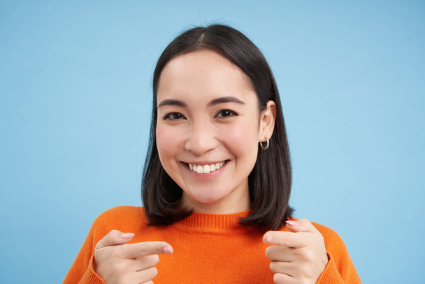 Gotcha. Nahaufnahme Porträt einer lächelnden asiatischen Frau zeigt zufrieden mit den Fingern in die Kamera, gratuliert, steht vor blauem Hintergrund. - Foto, Bild