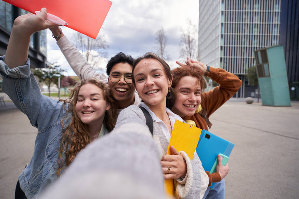Uśmiechnięci wielorasowi studenci robią sobie selfie z powrotem do szkoły. Szczęśliwi przyjaciele robią zdjęcia przed kampusem, trzymając foldery. Wesołe koleżanki i koledzy z klasy pozują i bawiają się.  - Zdjęcie, obraz