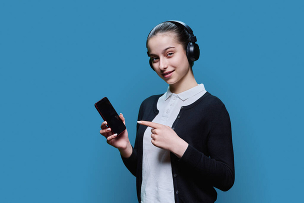 Young female in headphones listening audio watching video on smartphone, blue background. Teenage girl student studying resting. Adolescence, internet technology online, services for learning leisure - Foto, Bild