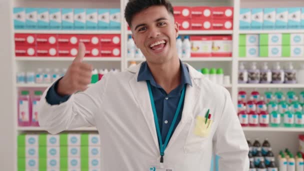 Jonge Spaanse man apotheker glimlachend zelfverzekerd doen ok gebaar met duimen omhoog bij apotheek - Video
