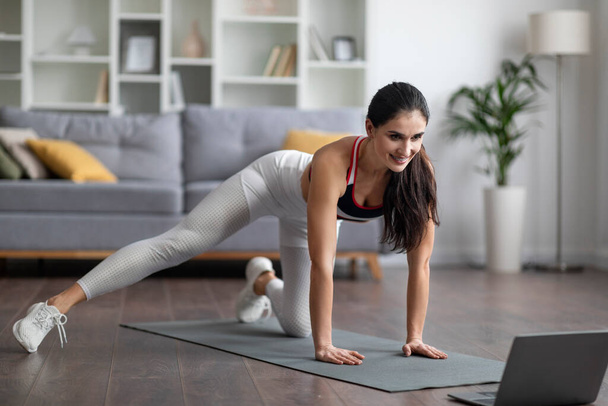Entraînement en ligne. Athlétique bien ajusté belle brunette Moyen-Orient jeune femme en vêtements de sport faisant pilates sur tapis de yoga à la maison, étirement des jambes, regarder la vidéo de remise en forme sur ordinateur portable PC, copier l'espace - Photo, image