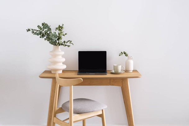 ανοιγμένο φορητό υπολογιστή, κεραμικό βάζο με κλαδί ευκαλύπτου και φλιτζάνι καφέ σε ξύλινο γραφείο κοντά σε άνετη καρέκλα σε λευκό φόντο τοίχου σε κομψό διαμέρισμα - Φωτογραφία, εικόνα