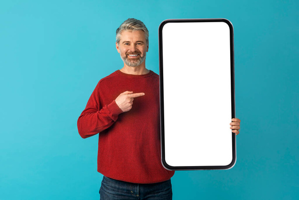 Schöner kühl lächelnder Mann mittleren Alters in lässiger Haltung mit riesigem nagelneuem Handy mit weißem Bildschirm in der Hand, posiert über blauem Studiohintergrund und zeigt auf eine Attrappe - Foto, Bild