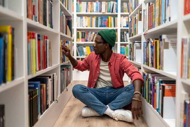 Studente pensieroso afro-americano che sceglie libri di ricerca nella biblioteca universitaria che li prende dalla libreria. Ragazzo premuroso hipster seduto sul pavimento a studiare in luogo pubblico. Istruzione, apprendimento. - Foto, immagini